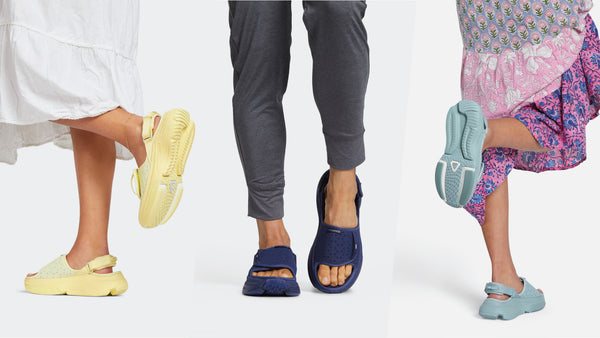 Three people wearing different colored thick platform slide sandals, shown from the waist down against a light gray background.